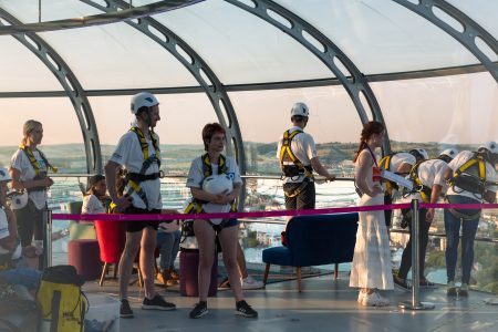 A group of abseil participants with safety gear inside a curved glass structure overlooking a scenic landscape at sunset.