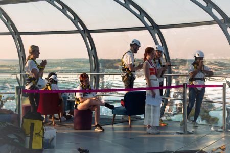 Charity event participants in safety gear inside a glass dome structure with panoramic views at sunset.