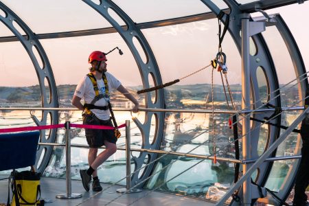 A participant in a red helmet and safety harness looks out over the cityscape at a charity abseil event during sunset.