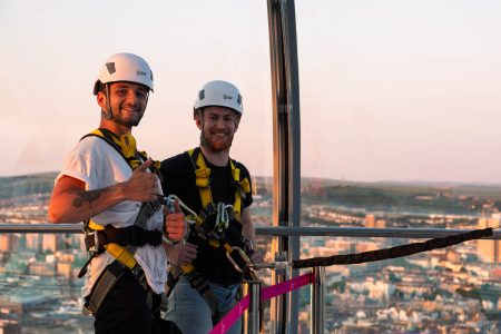 Two cheerful men in abseiling gear giving a thumbs-up before a charity abseil with a cityscape backdrop at sunset.