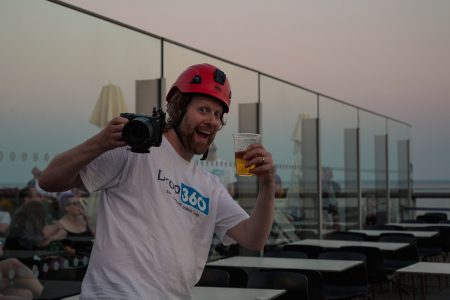 Smiling man with a red helmet holding a DSLR camera in one hand and a beer in the other, at a rooftop bar during dusk