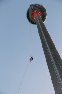 Person descending from a tall tower on a zip line at dusk.