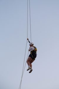 Adventurous individual zip-lining against a clear sky, with safety gear and helmet on.