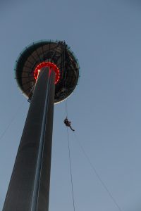 A person zip-lining from a tall tower with a glowing red ring at the top in the twilight sky