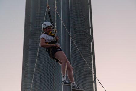 A person in a harness smiling while descending a zip line against a modern tower at dusk.