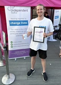 Smiling man with a certificate and mug in front of Independent Lives charity banner.