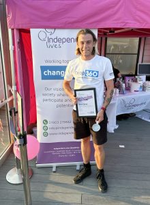 A man with long hair smiling while holding a certificate and mug in front of an Independent Lives charity stand.
