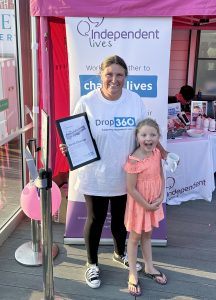 A smiling woman and a child holding a certificate and mug standing in front of an "Independent Lives" charity banner.