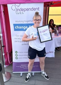 A woman stands proudly with a certificate and mug in front of an Independent Lives charity stand.