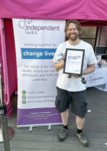 A man stands smiling, holding a certificate and a mug in front of a charity banner for Independent Lives.