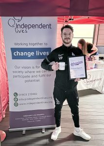 A man proudly holds a certificate and a mug in front of an Independent Lives charity banner.