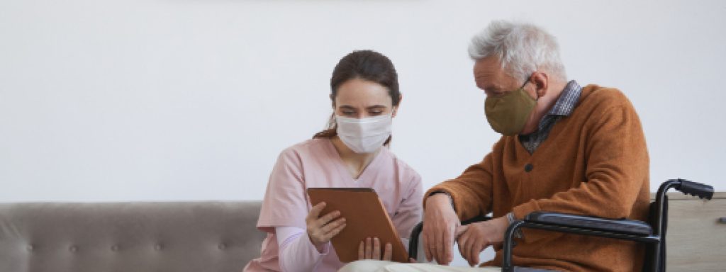 An elderly man in a wheelchair and a woman wearing face masks while looking at a tablet together