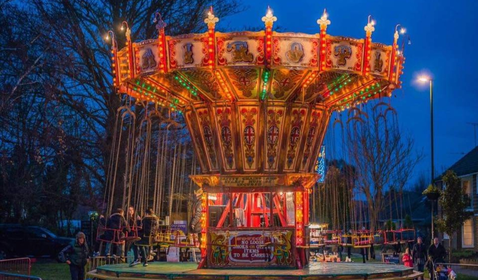 An illuminated vintage carousel at twilight with people nearby in a festive setting