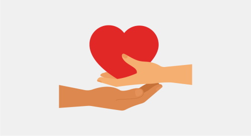 Two hands of different skin tones, one from above and one from below, engaged in a caring gesture. The hand from above is holding a vibrant red heart and is gently placing it on the palm of the hand below, symbolizing giving, compassion, and support. The background is plain and light-coloured, putting the focus on the central act of kindness.