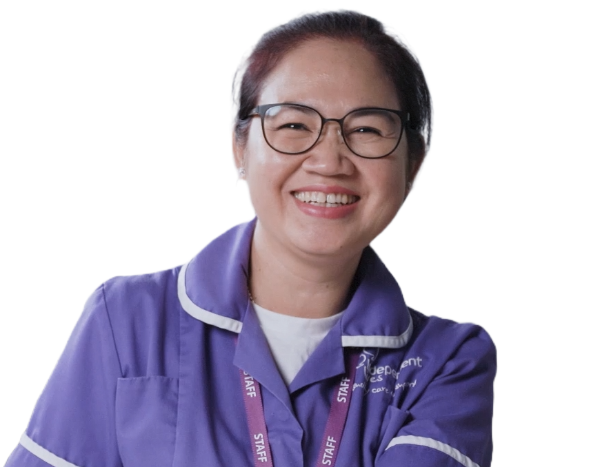 The image depicts a cheerful middle-aged woman with glasses, wearing a purple uniform with a visible name tag that reads "STAFF". Her hair is short and neatly styled, and she's sporting a broad, friendly smile. The uniform suggests she may be an employee, possibly in a service or care role. The background of the photo is transparent, focusing attention solely on the individual.