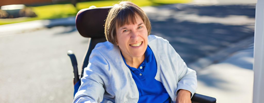 A smiling woman with short hair in a wheelchair, wearing a blue shirt and light jacket outdoors