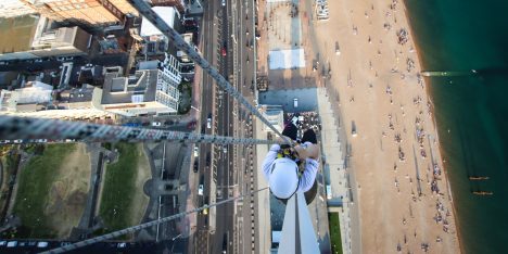A bird's-eye view of an individual rappelling down a high building with a coastal road and beach visible below.