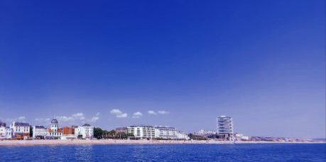 A serene seascape with a clear blue sky above and calm waters below. The coastline is dotted with buildings, including a distinctive structure with a domed roof.