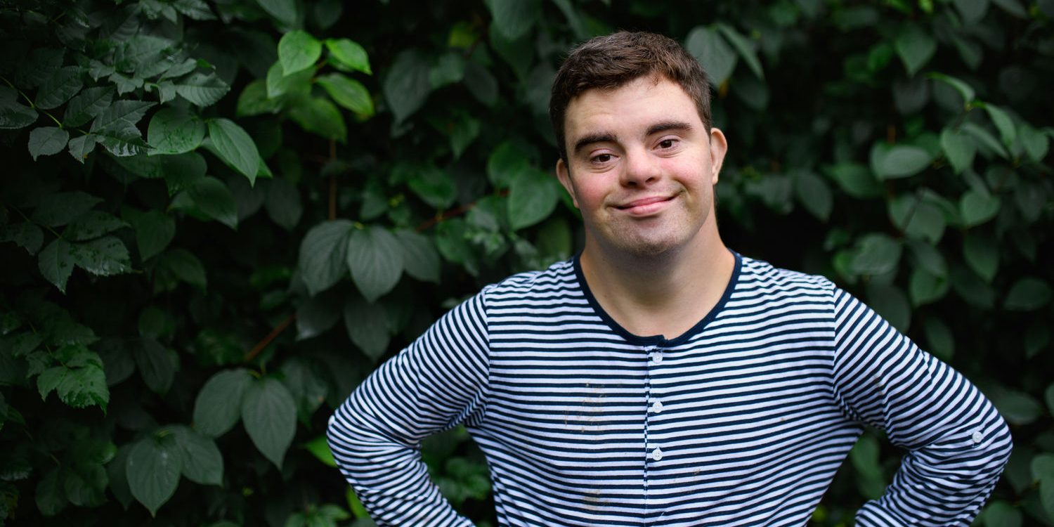 A young man with Down syndrome stands confidently in front of a lush green hedge, smiling in a striped shirt.