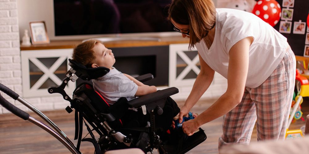 A caring adult secures a child's harness in a modern wheelchair within a cozy home setting.