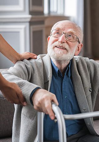 Elderly man with glasses smiling upwards at a caregiver in blue attire, holding onto a walking frame.