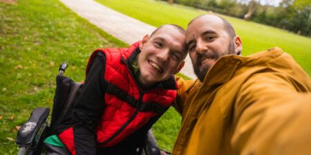 Two friends, one in a wheelchair, taking a selfie together in a park.