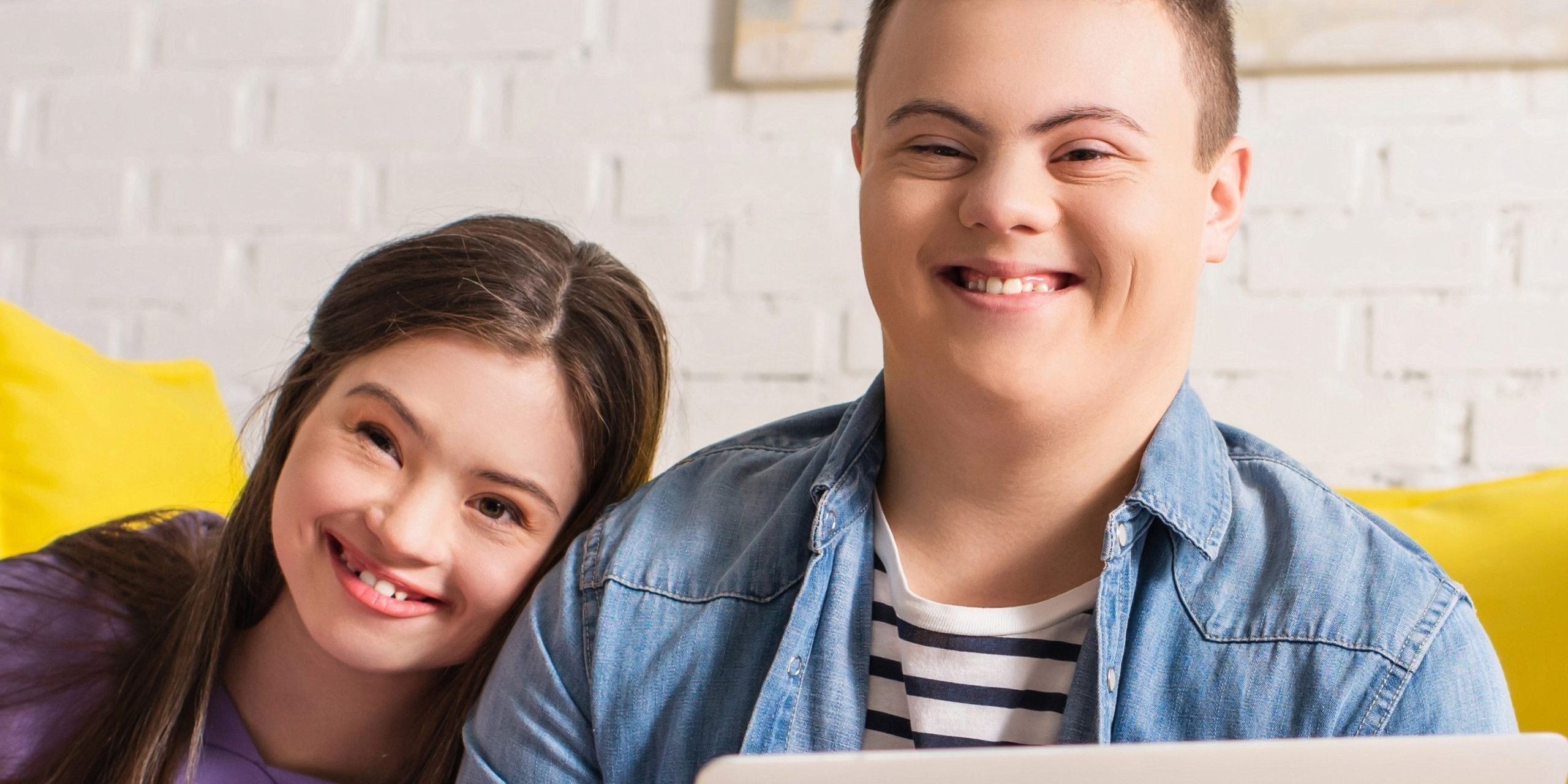 A young woman and a young man with Down syndrome smiling at the camera, sitting together with a laptop in a bright room with a white brick wall in the background.