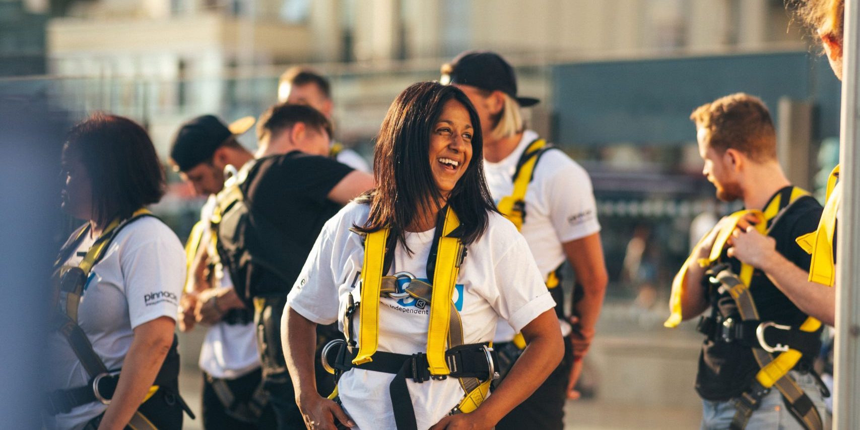 A smiling woman in a harness stands among a group of people preparing for an adventurous activity.