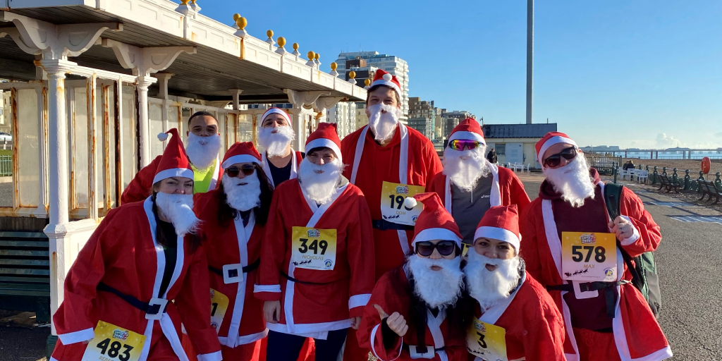 The image shows a group of people dressed in Father Christmas costumes, complete with red hats, jackets, and white beards. They are wearing numbers, suggesting they are participating in a themed fun run or charity race. They're posing for the photo with a backdrop of a classic seaside promenade, identifiable by the white balustrade and shelter, under a clear blue sky. The setting appears to be a bright, sunny day, likely along a coastal area, possibly during a festive season event.