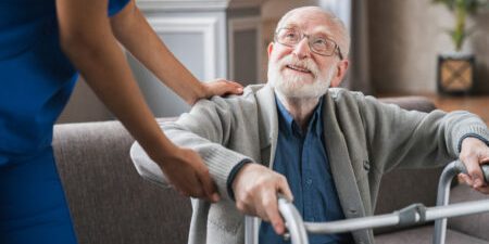 Elderly man with glasses smiling upwards at a caregiver in blue attire, holding onto a walking frame.