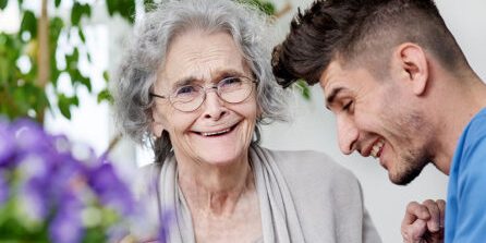 An elderly woman with glasses smiling broadly as a young man leans in close, sharing a joyful moment.