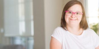 Smiling woman with Down syndrome wearing pink glasses, posing indoors.