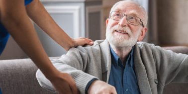 Elderly man with glasses smiling upwards at a caregiver in blue attire, holding onto a walking frame.