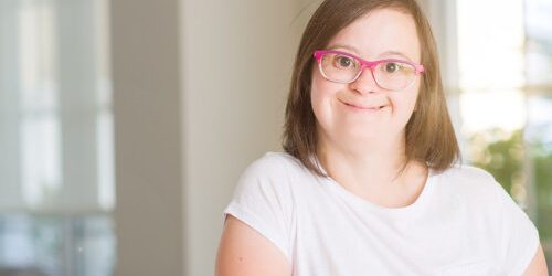 Smiling woman with Down syndrome wearing pink glasses, posing indoors.