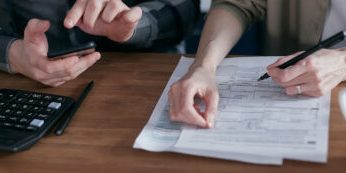 Two people collaborating on paperwork with a calculator and smartphone on a wooden table.