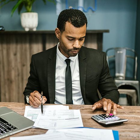 A focused businessman in a black suit meticulously reviews financial documents at his desk with a calculator and laptop.