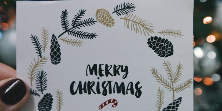 Hand holding a Christmas card with 'Merry Christmas' surrounded by pine illustrations, against a blurred tree backdrop.