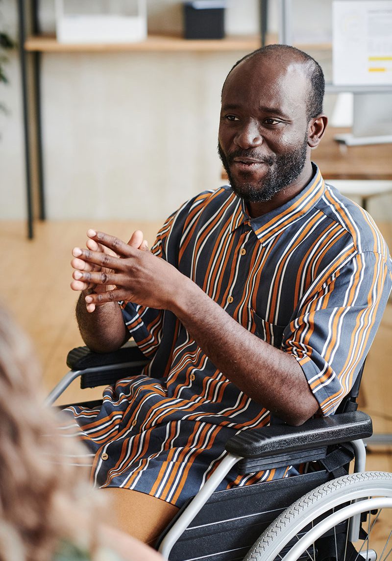 A man in a striped shirt smiling and clapping hands while seated in a wheelchair.
