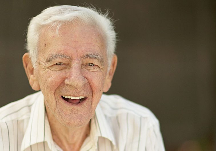 Elderly man smiling broadly in a light striped shirt with a dark background.