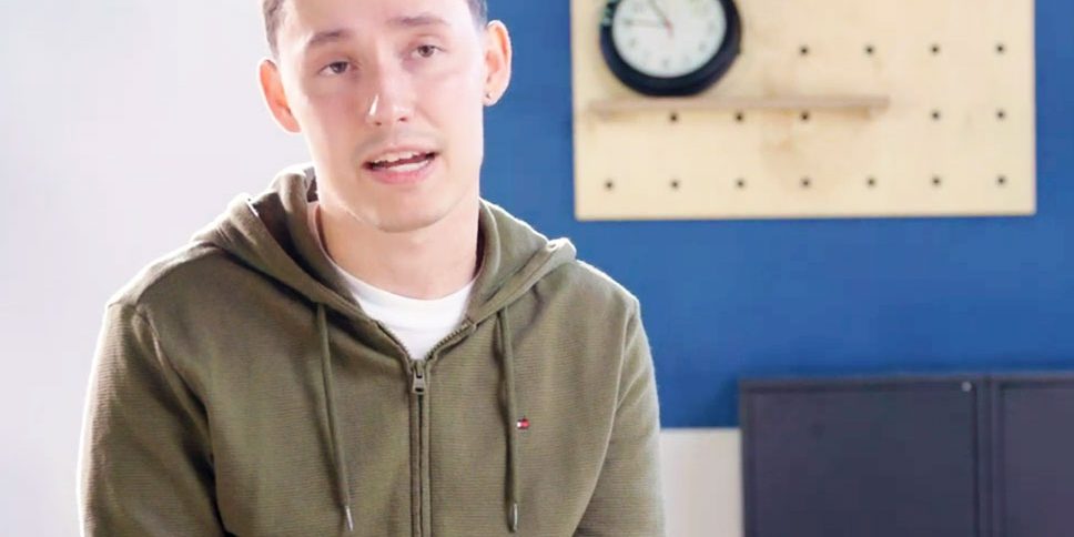 Young man in a green hoodie sitting indoors with a blue wall and clock in the background.