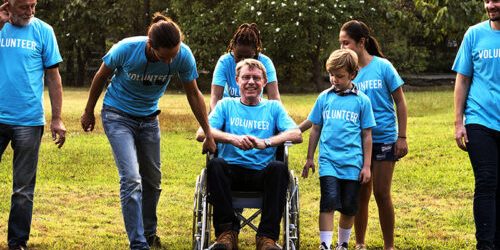 A diverse group of people wearing blue 'VOLUNTEER' t-shirts outdoors, including a person in a wheelchair.